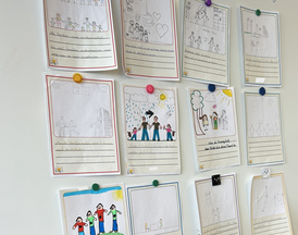 Kartenentwürfe die auf einer Tafel hängen. Es sind 12 Kartenentwürfe zu sehen. Es sind A4 Zettel auf denen Kinder in ein Feld eine Zeichnung zu dem darunter stehenden kurzen Satz gemalt haben. 