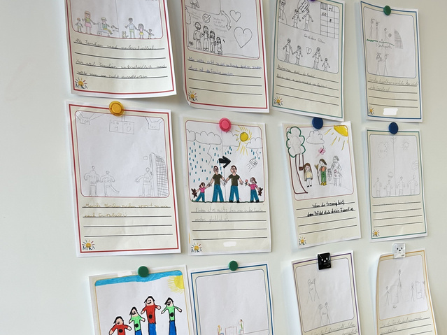Kartenentwürfe die auf einer Tafel hängen. Es sind 12 Kartenentwürfe zu sehen. Es sind A4 Zettel auf denen Kinder in ein Feld eine Zeichnung zu dem darunter stehenden kurzen Satz gemalt haben. 